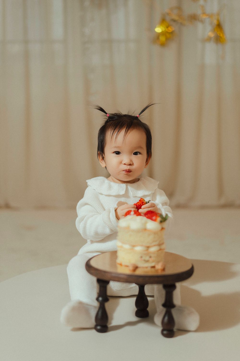 Small child in white outfit sits with mini cake on wooden pedestal during photo session with gold decorations.