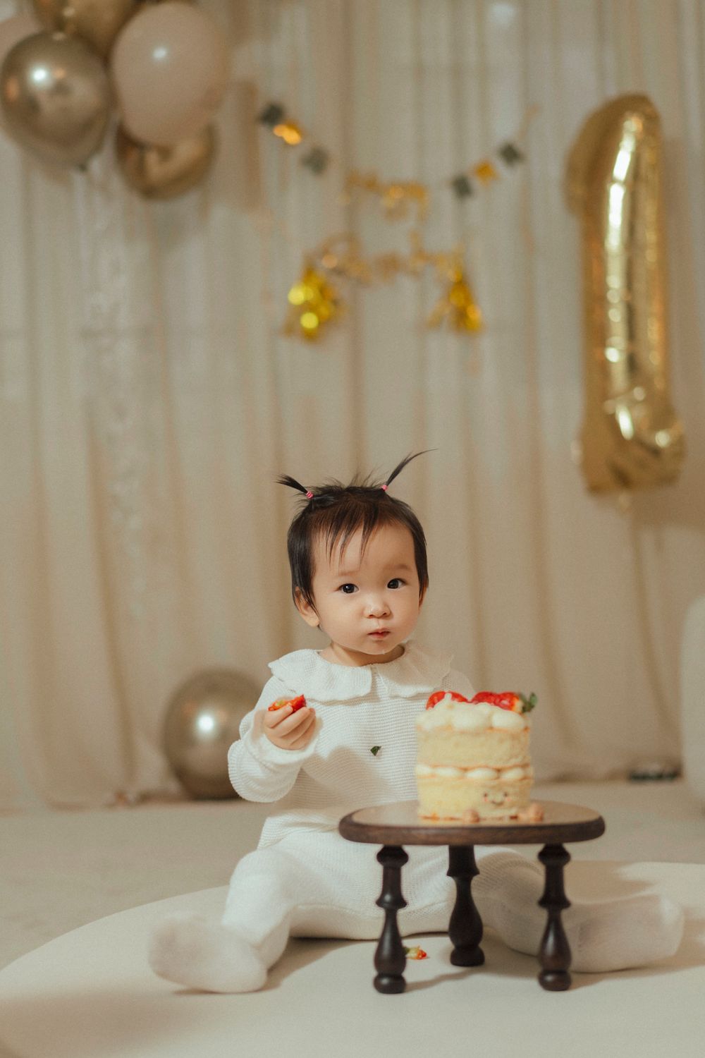 A small child in white clothing sits beside a birthday cake with gold balloons and decorations in the background.