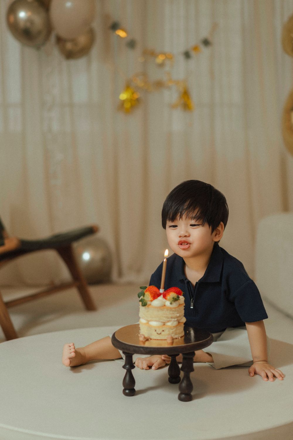 A child sits at a white table with a decorated fruit cake on a black pedestal stand while balloons decorate the background.