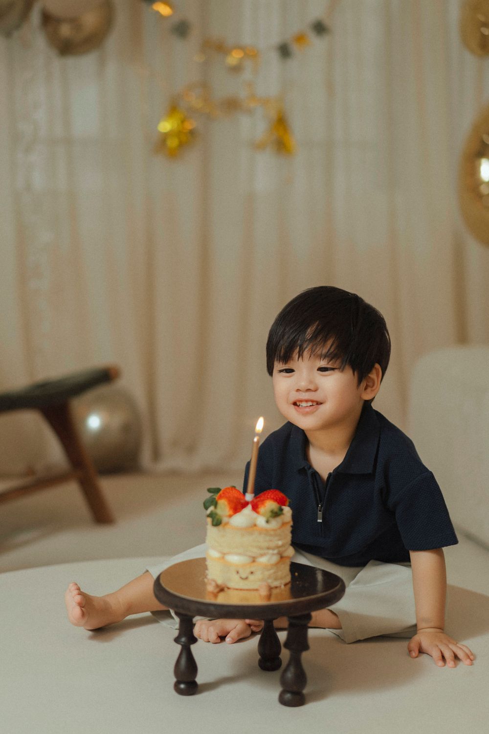 A decorated birthday cake sits on a dark wooden stand while soft lights and party decorations glow in the background.
