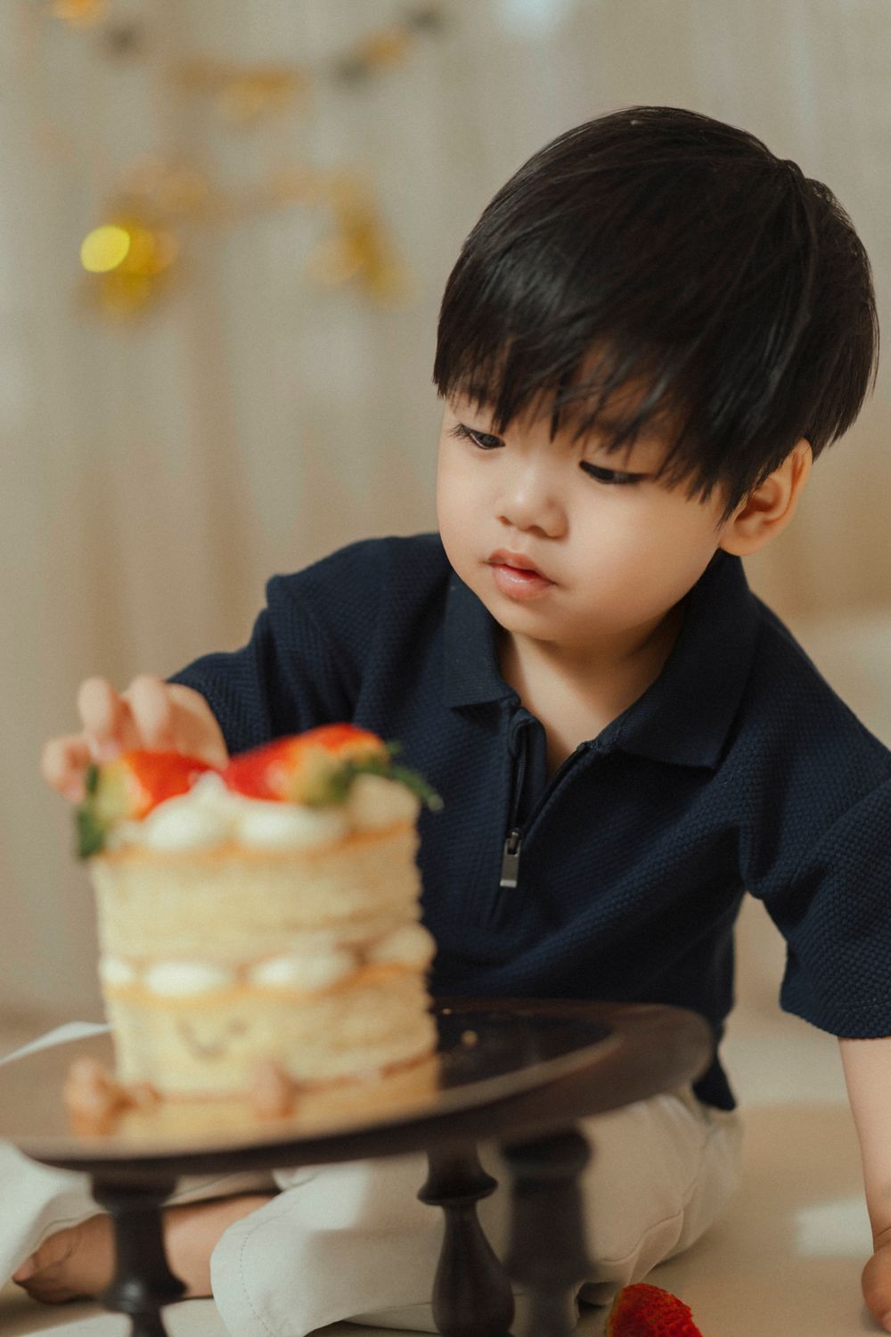 A child in a navy polo shirt gazes at a layered vanilla cake topped with strawberries on a black pedestal stand.
