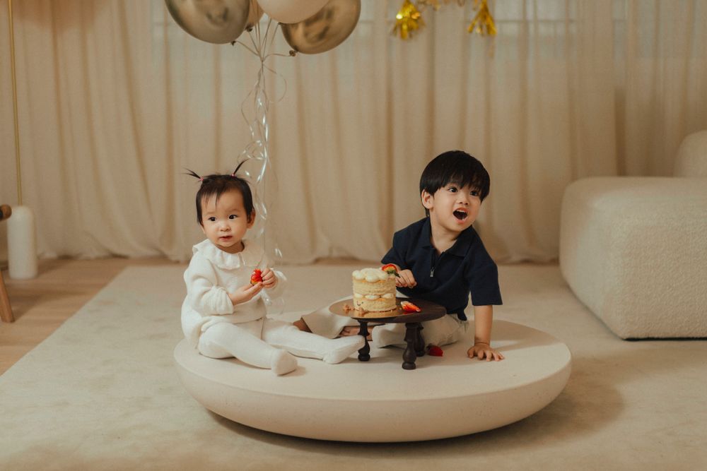 Two young children sit on a round platform with balloons and a small cake display in a neutral-toned room.