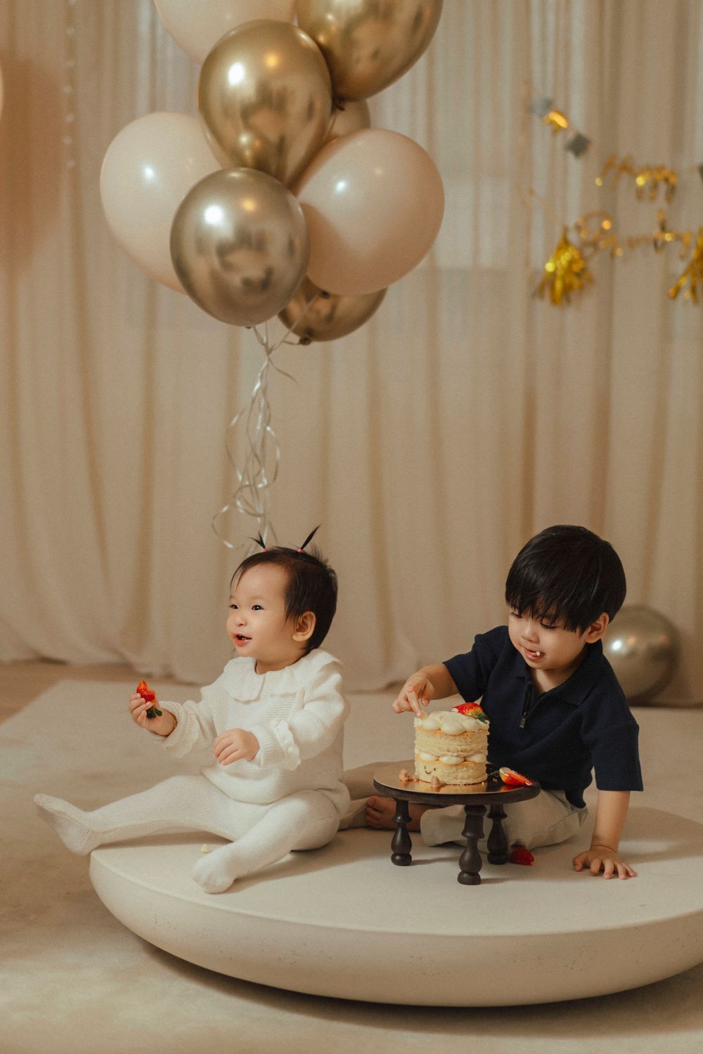 Two children sit on a round white platform with gold and white balloons and a small cake for a celebration.