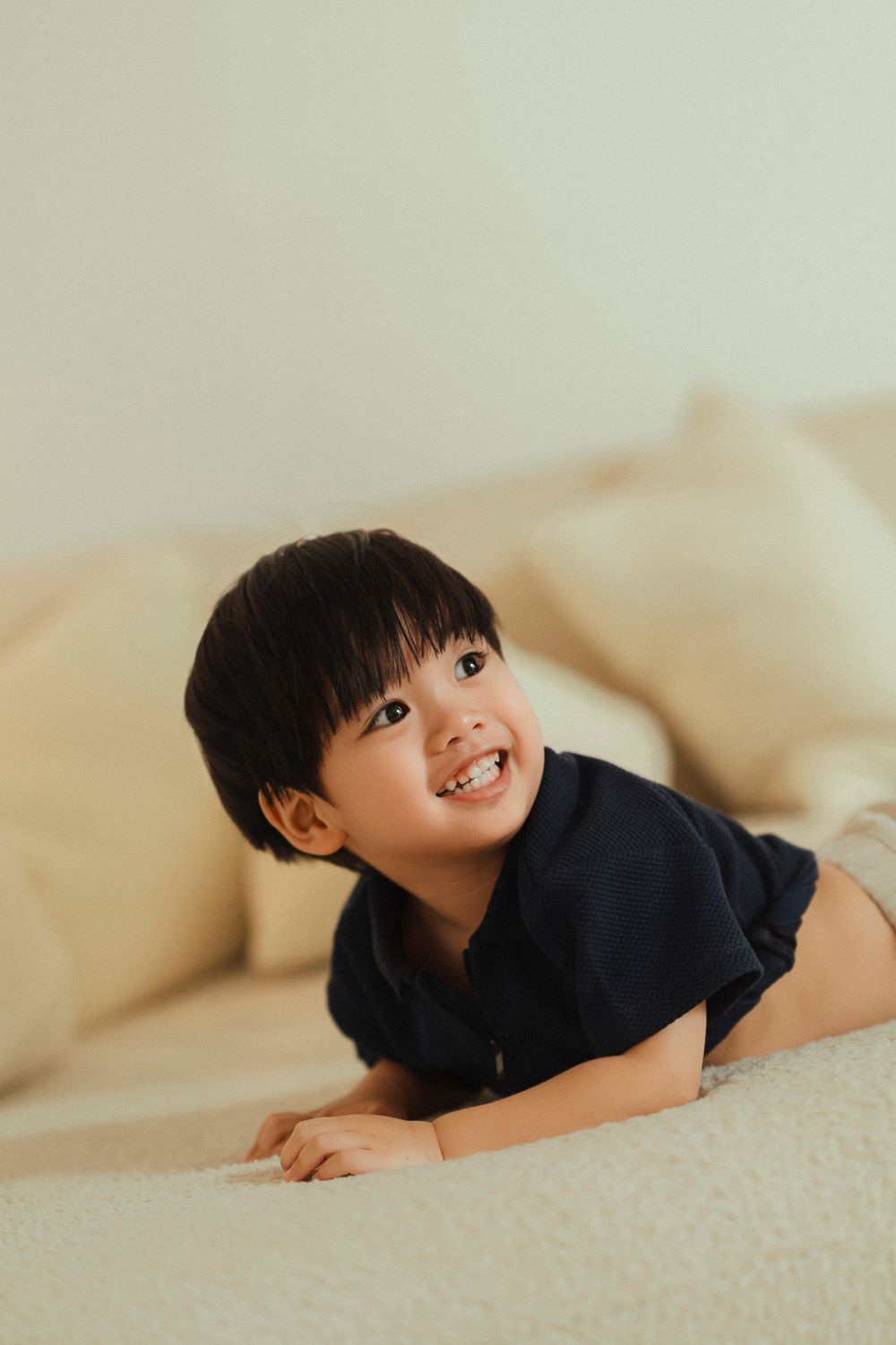 Smiling child in a navy blue shirt lying on a beige couch in a warmly lit room.
