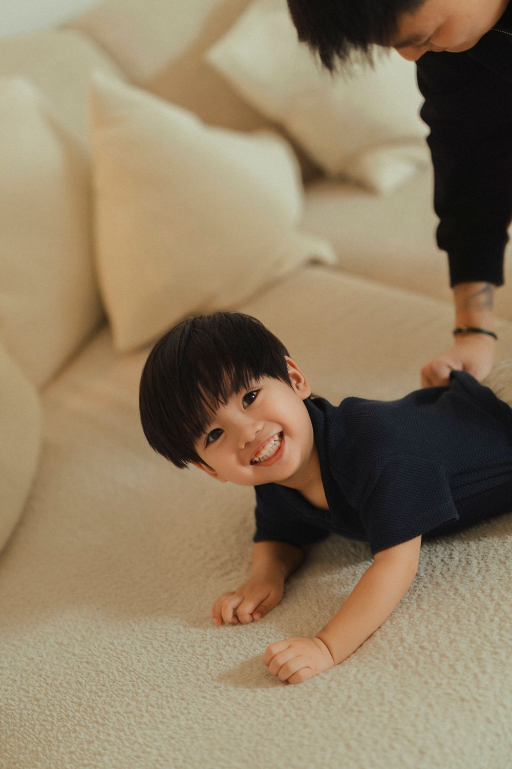 A young child in a navy blue shirt lies playfully on a beige carpet near a light-colored couch.