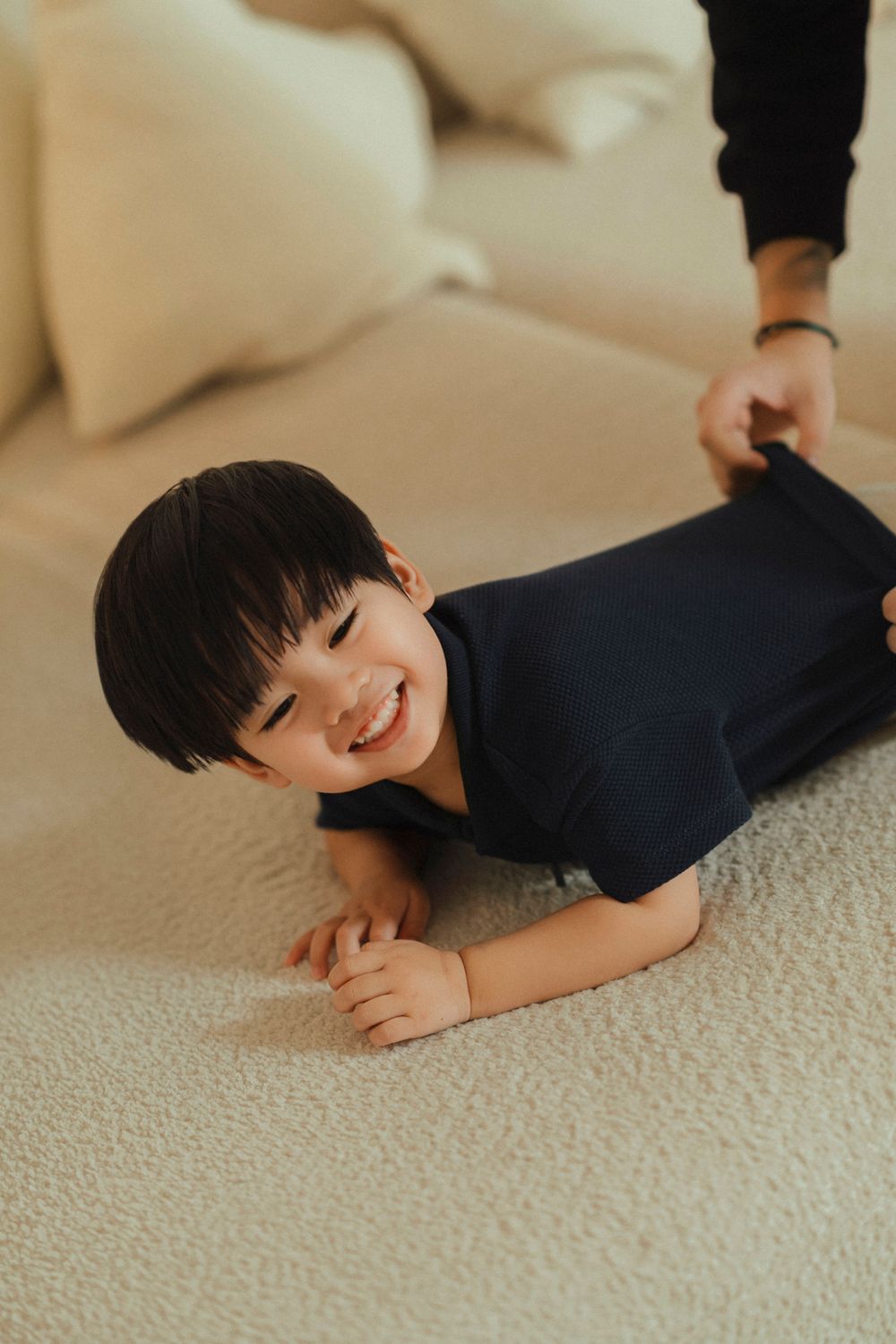 Child wearing navy blue outfit lies on beige carpet with a bright smile while playing on the floor.