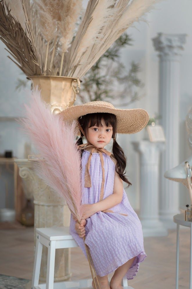 Figure in purple dress holds pink pampas grass while sitting on white chair in elegant setting.