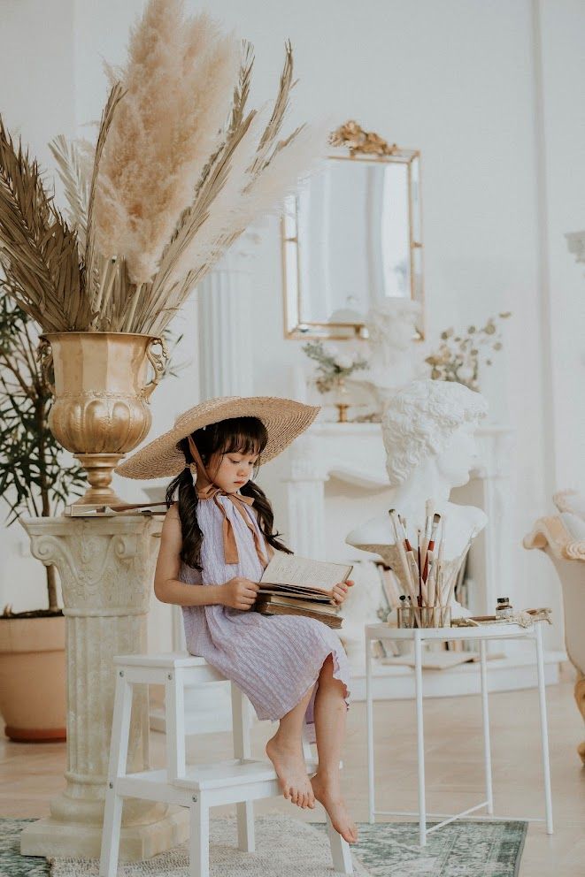 Figure in purple dress and straw hat reads book while sitting in decorated white room with pampas grass.