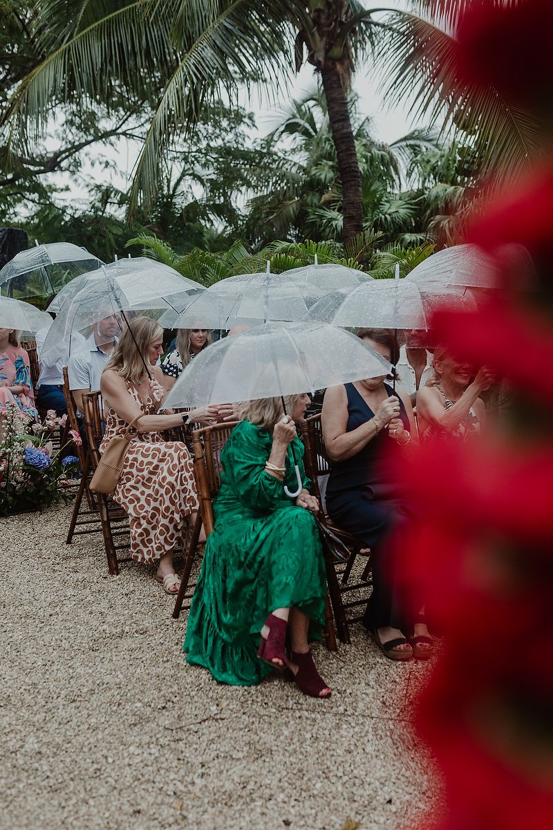 Guests holding clear bubble umbrellas at an outdoor tropical wedding ceremony on a rainy day.
