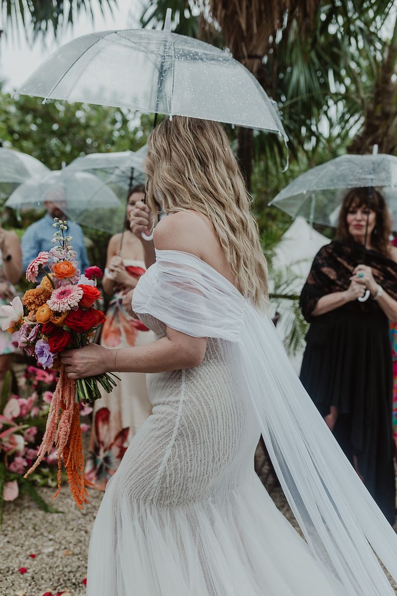 Bride in white off-shoulder gown holds colorful bouquet under clear umbrella at outdoor rainy wedding ceremony.