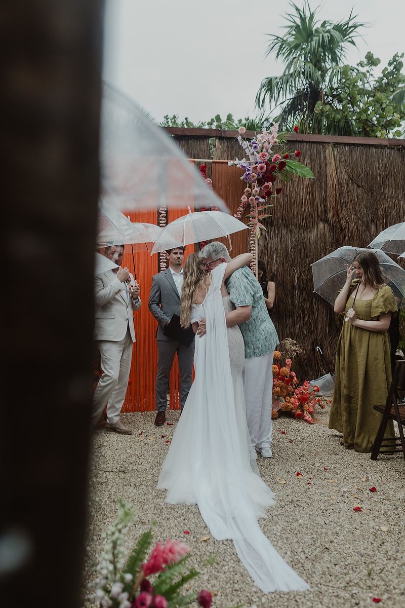 Bride and groom exchange vows outdoors in the rain, guests holding clear umbrellas at a boho wedding ceremony.