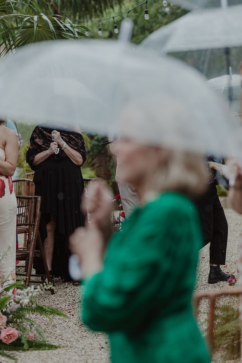 Guests holding clear umbrellas at an outdoor rainy wedding ceremony, woman in green jacket in foreground.