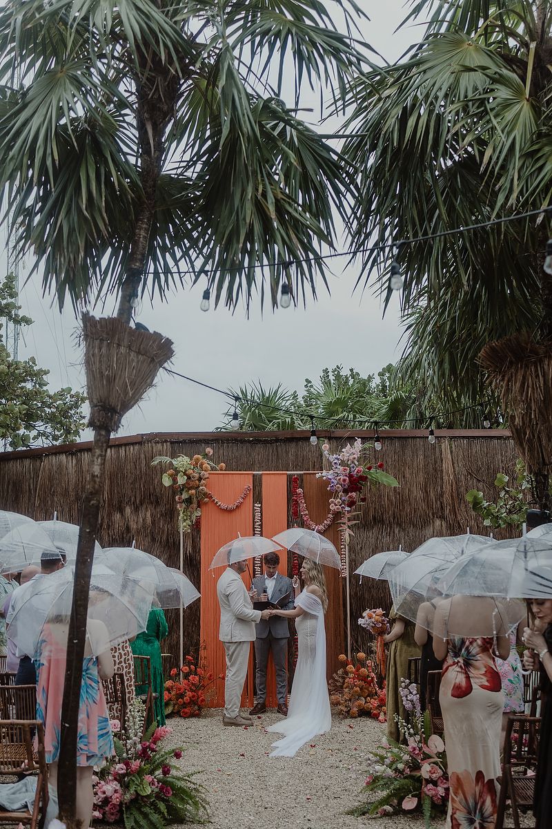 Outdoor wedding ceremony in the rain with guests holding white umbrellas, tropical palm trees, and colorful floral arch backdrop.