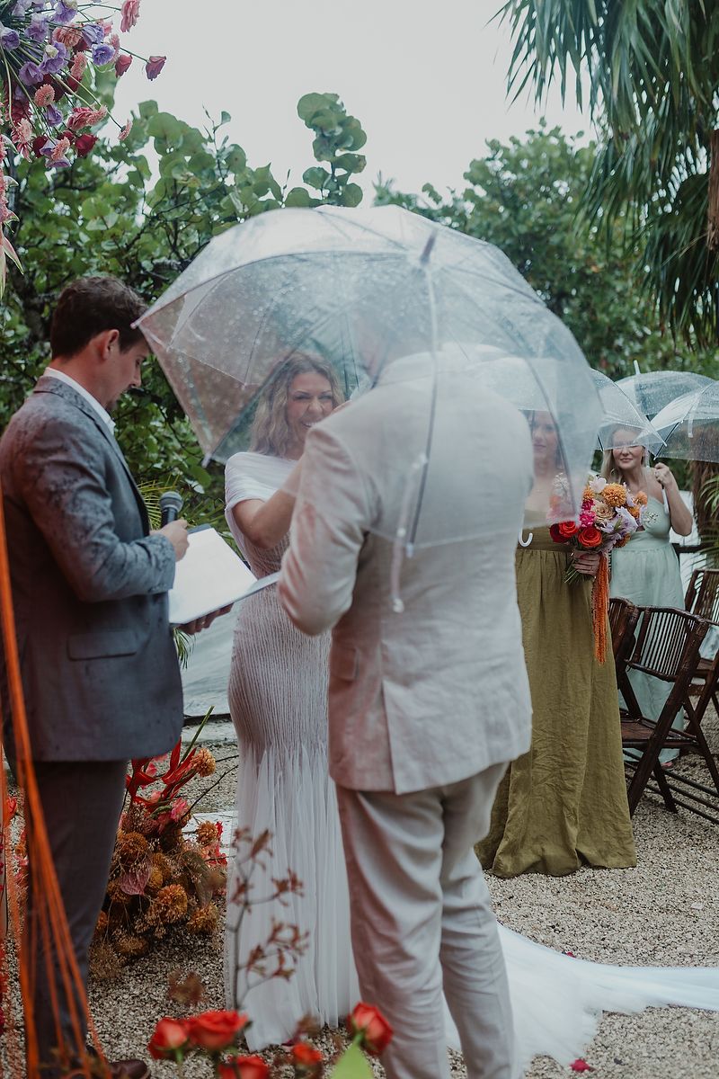 Bride and groom exchange vows under clear umbrellas during a rainy outdoor wedding ceremony surrounded by colorful floral arrangements.