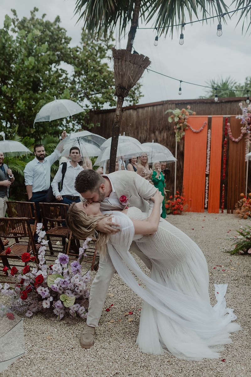 Groom dips bride for a romantic kiss at an outdoor wedding ceremony with guests holding clear umbrellas.