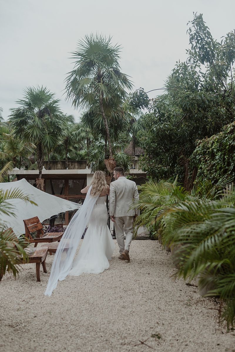 Bride and groom walk hand-in-hand through a tropical garden path lined with palm trees on their wedding day.