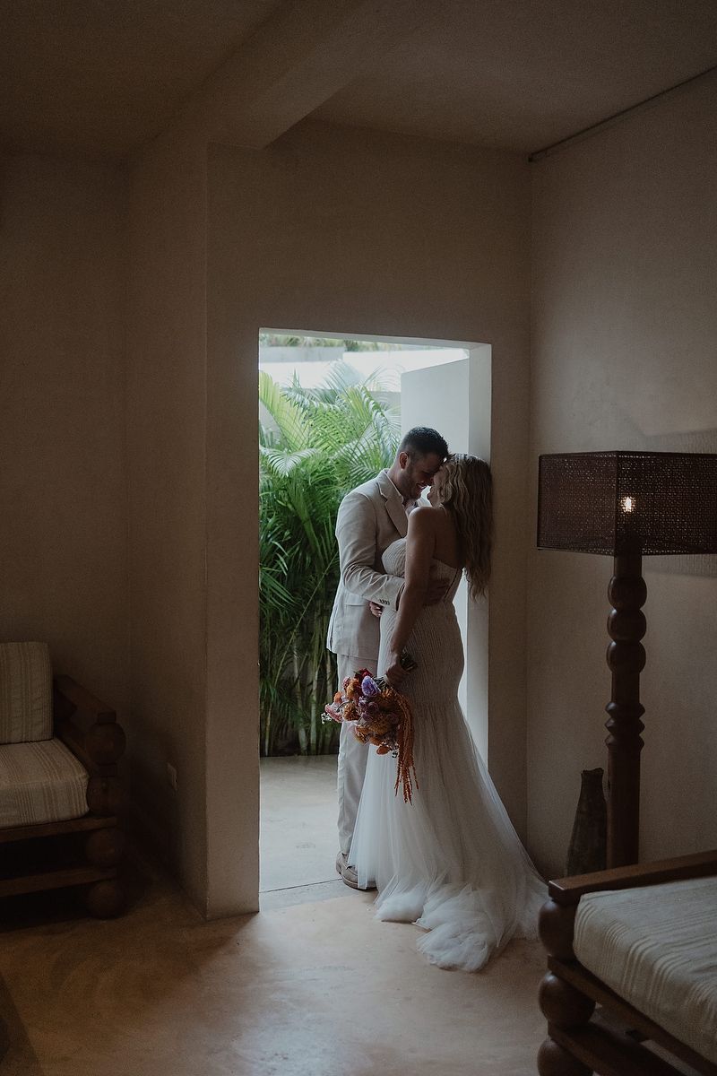 Bride and groom sharing a romantic kiss in a doorway, bride holding a colorful bouquet in a tropical wedding setting.