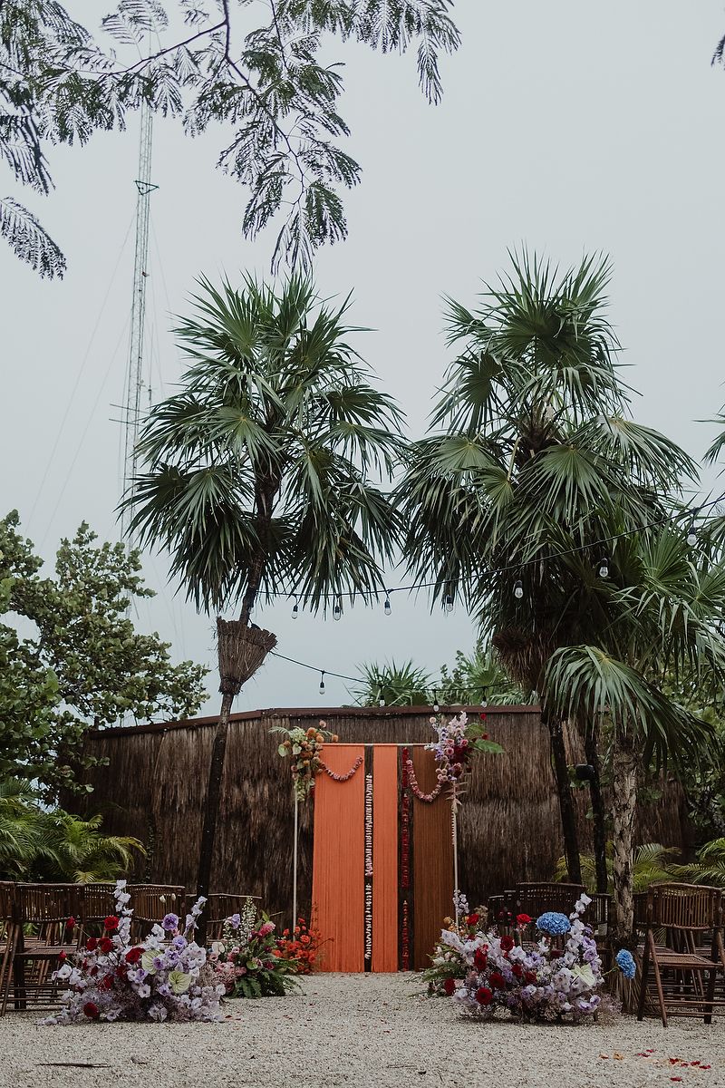 Outdoor wedding ceremony setup with coral arch, floral arrangements, palm trees, string lights, and wooden chairs on sandy ground.