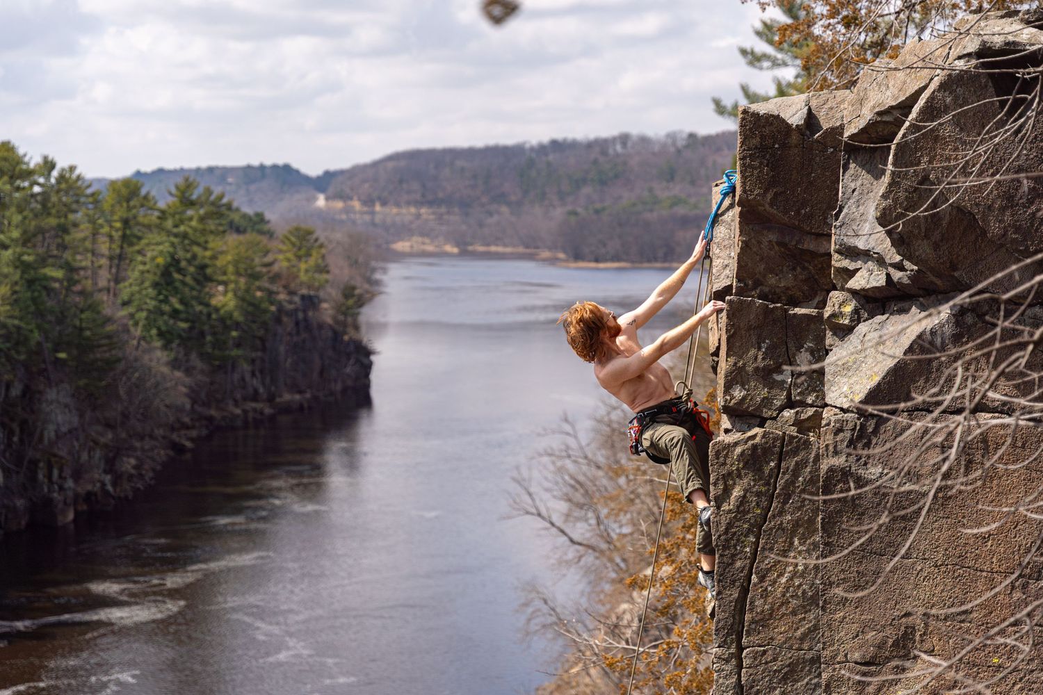 A rock climber scales a steep cliff face overlooking a serene lake surrounded by pine forests at the Interstate State Park.