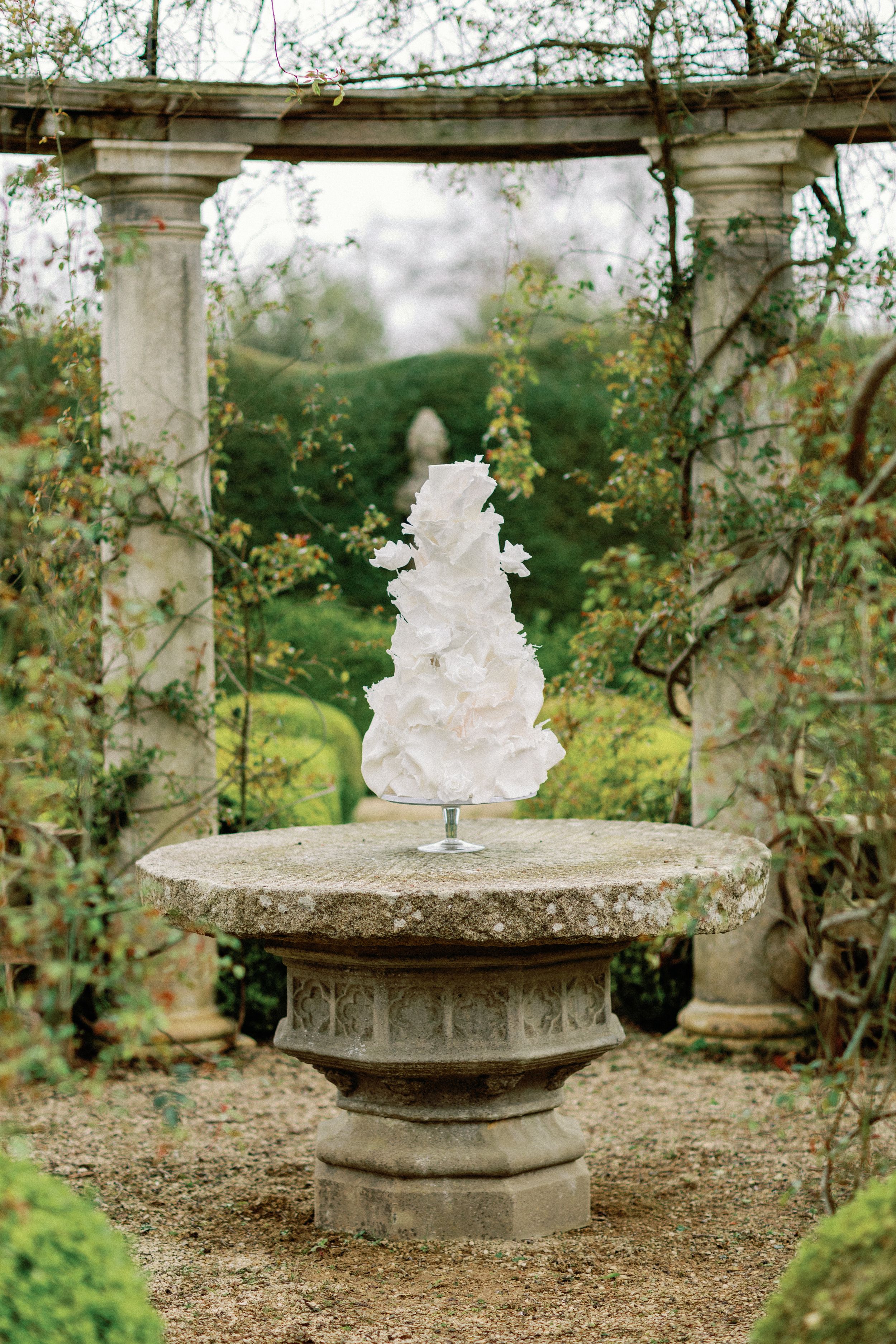 Elegant white wedding cake displayed on stone pedestal in garden pergola.