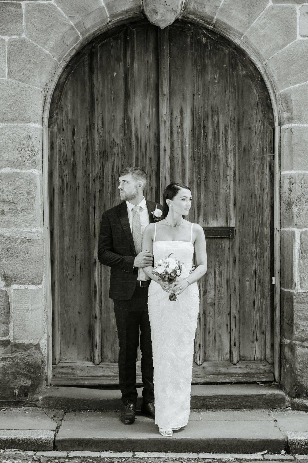 Artistic black and white wedding portrait under stone archway with wooden door.