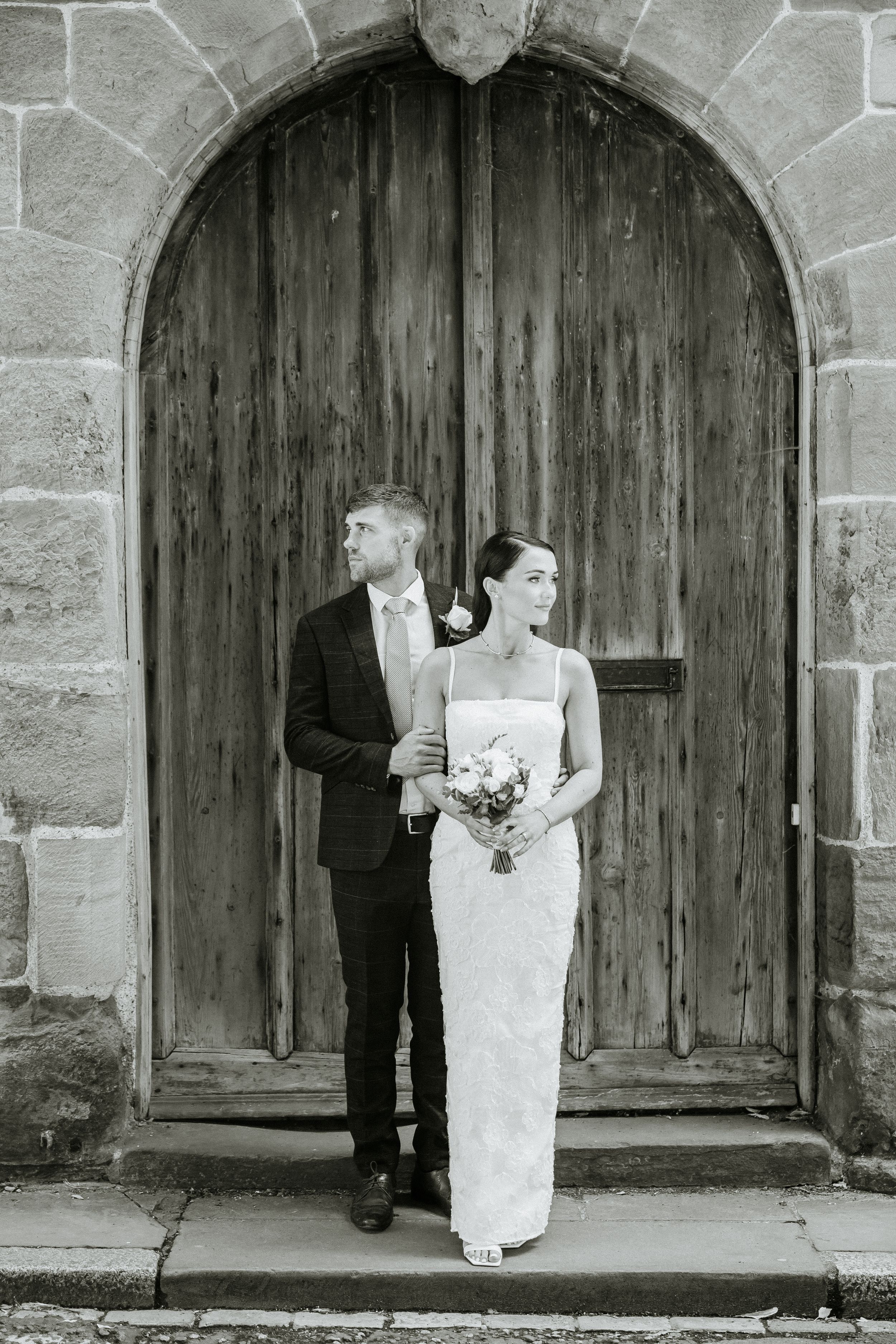 Artistic black and white wedding portrait under stone archway with wooden door.