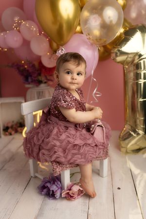 Baby in a dusty rose sequined dress sits on white wooden floor surrounded by pink and gold balloons.