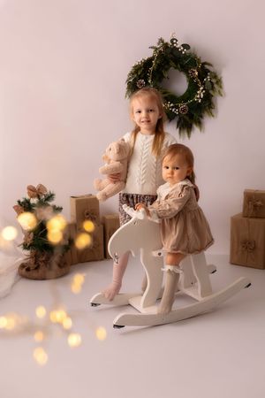 Children in cozy winter outfits sit on rocking horses with holiday decorations and warm glowing lights in the background.