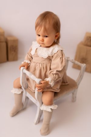 A baby in a beige vintage-style dress and white collar sits on a white surface in various poses.