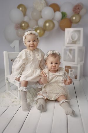 Twin babies in matching white vintage outfits posing with decorative letters and balloons.