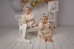Studio portrait of babies in white dresses with number one prop and balloon decorations.