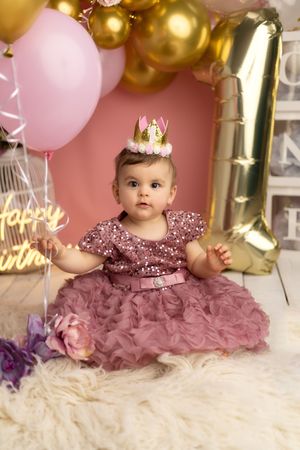 First birthday celebration setup with pink and gold balloons, sparkly dress, and tiny crown against pink backdrop.