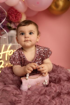 A baby in a sparkly pink tulle dress poses during a professional photo session with balloons and decorations.