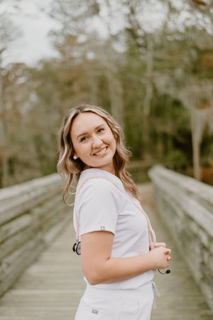 A person in white scrubs smiles while standing on a wooden boardwalk surrounded by blurred trees in the background.