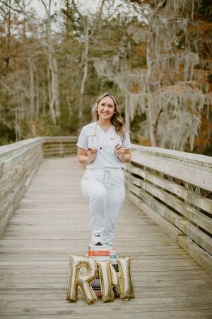 Healthcare worker in scrubs celebrates graduation with gold letter balloons on a wooden boardwalk surrounded by trees.