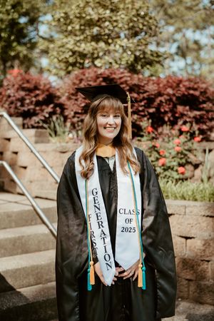 Graduate in black cap and gown with white sash stands on stone steps with red and green foliage in background.