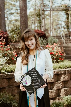 Graduate in white blazer holds decorated graduation cap while standing in garden with red flowers and stone wall.