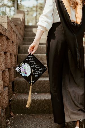 A hand holds a decorated black graduation cap with floral designs while standing on stone steps.