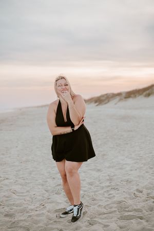 A person in a black dress stands smiling on a sandy beach during sunset.