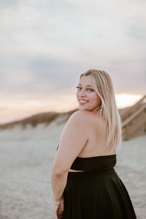 A person in a black strapless dress poses against sandy dunes at sunset with a dreamy coastal backdrop.