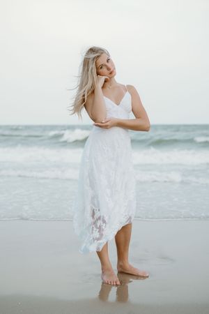 Blonde woman in flowing white lace dress standing barefoot on ocean shoreline with wind in hair.