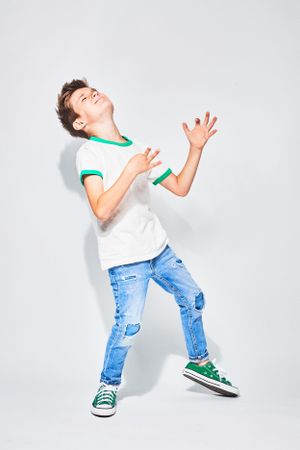 Boy in white T-shirt, ripped jeans, green sneakers, mid-dance with arms raised and a joyful expression