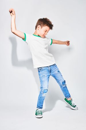 A young boy in a white T-shirt with green trim, ripped blue jeans, and green sneakers dances energetically. His head is tilted back with a j