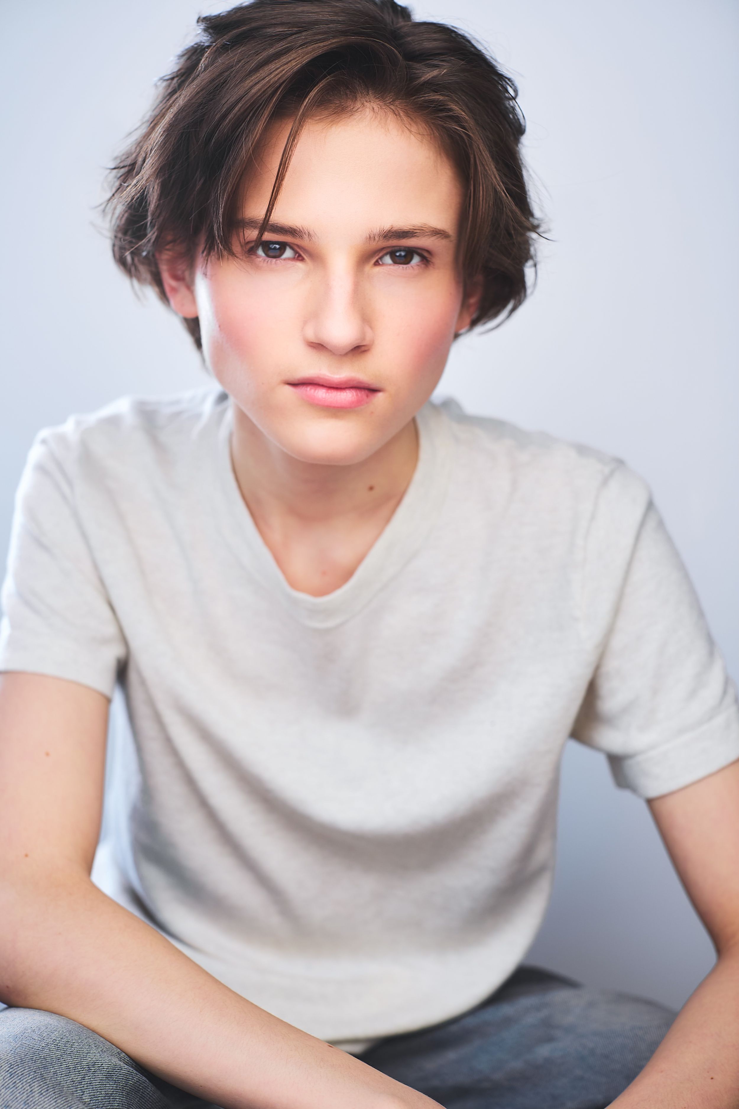 Headshots of a young boy with long hair in a white t-shirt sitting and leaning on his knees with a tendril of hair on his forehead.