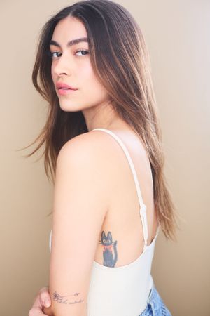 Portrait of a beautiful woman with brown hair in a white tank top turning to the side and showing a cat tattoo on her mid back.