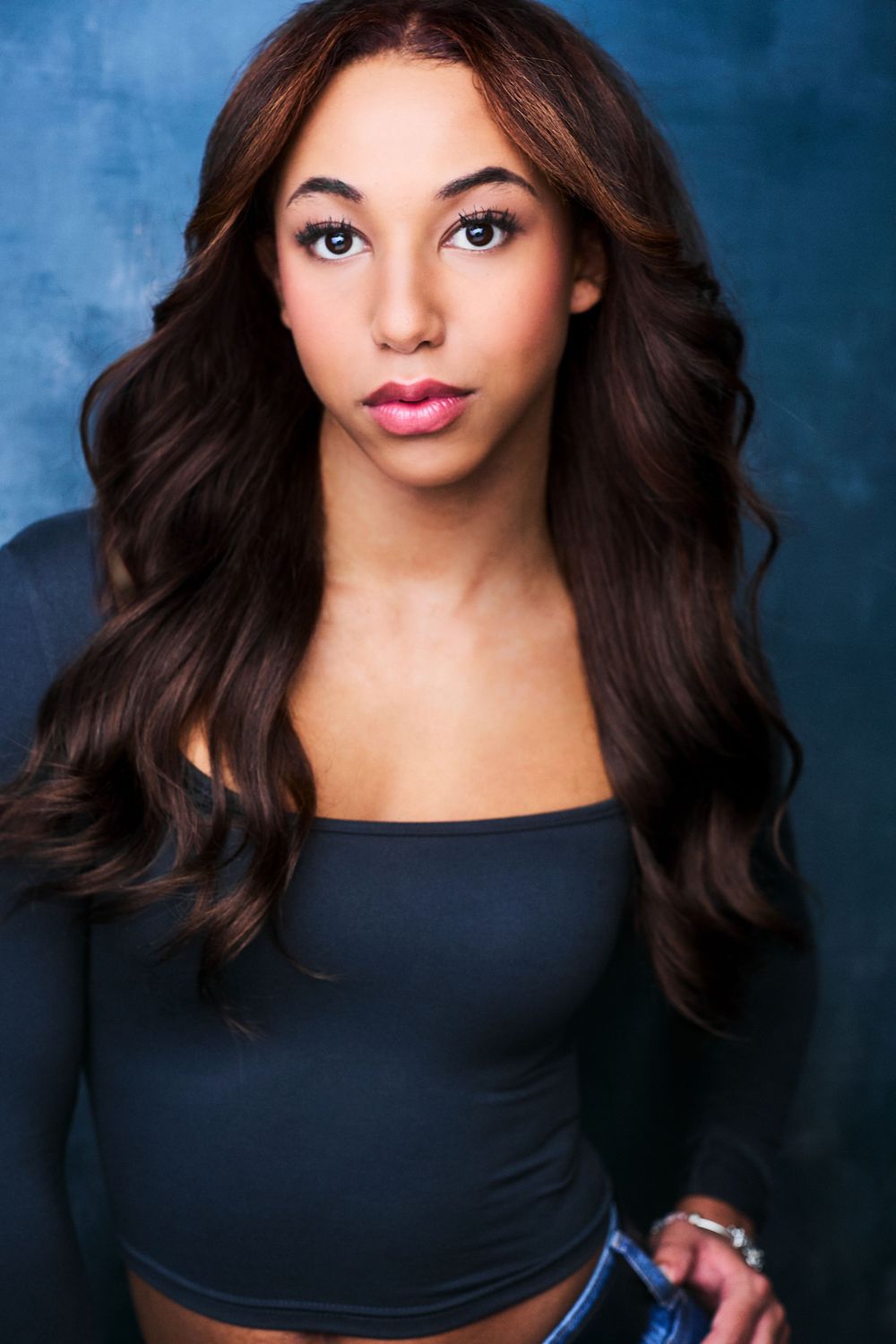 Headshot of a beautiful actor in a blue shirt with long brown hair against a blue textured background.
