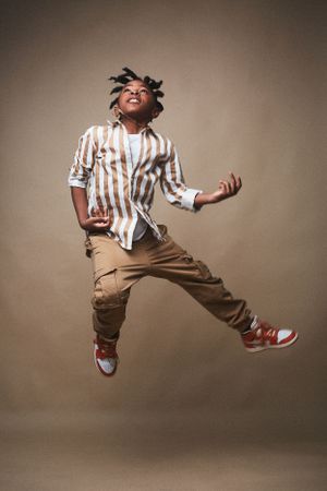 Headshot of a young boy with braids in a brown and white striped shirt suspended in the air in a joyful jump.