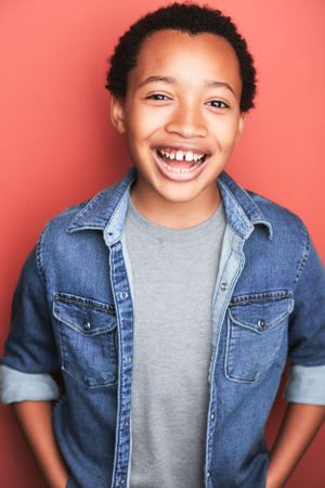 Headshot of an adorable young boy smiling brightly in a denim shirt against an orange background.