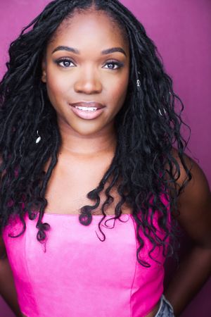 A woman with long braided hair wearing a pink top stands against a purple background, looking at the camera.