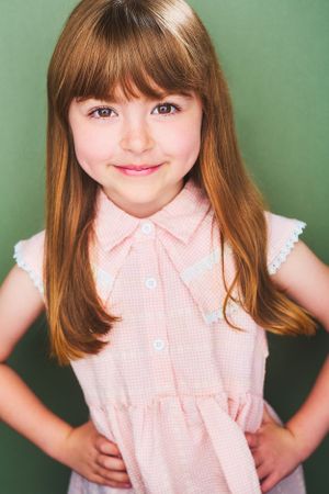 Headshot of an adorable young girl in a peach dress with red hair and her hands on her hips against a green background.